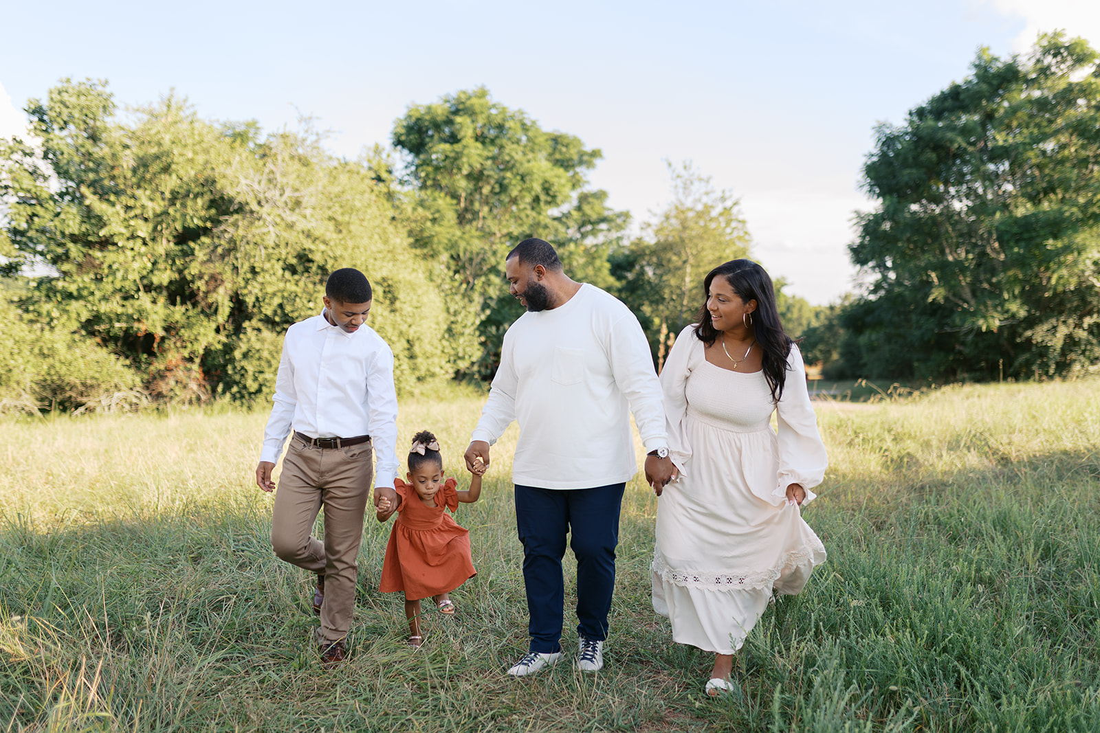 A family photography session at Little Mulberry Park in Dacula, GA. A family of four walks hand in hand through a grassy field under soft evening light, dressed in coordinating neutral and earthy tones. Captured by Atlanta family photographer Denay Shook