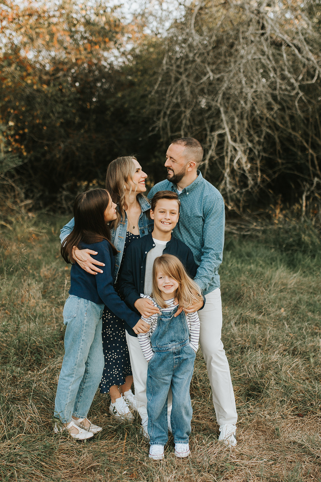 Family huddled together outdoors in coordinating denim-style outfits, smiling at each other during a lifestyle family photography session in Atlanta, Georgia. Captured by Denay Shook Photography, top-rated Atlanta family photographer.