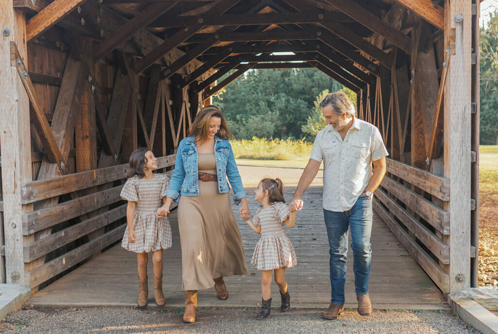 Family of four walking hand in hand across the covered bridge at Garrard Landing Park in Alpharetta, Georgia during golden hour, dressed in neutral Ralph Lauren-inspired fall outfits.