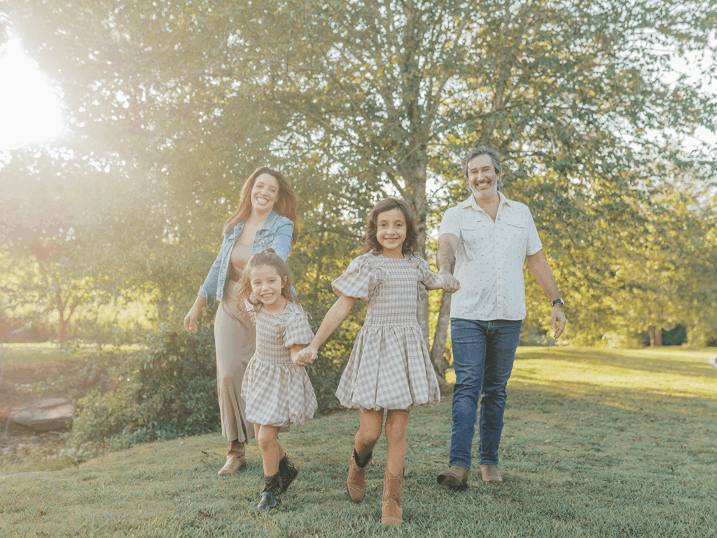 Smiling family of four walking through grassy field at sunset in Alpharetta, Georgia, with parents and two daughters in coordinated fall outfits at Garrard Landing Park.