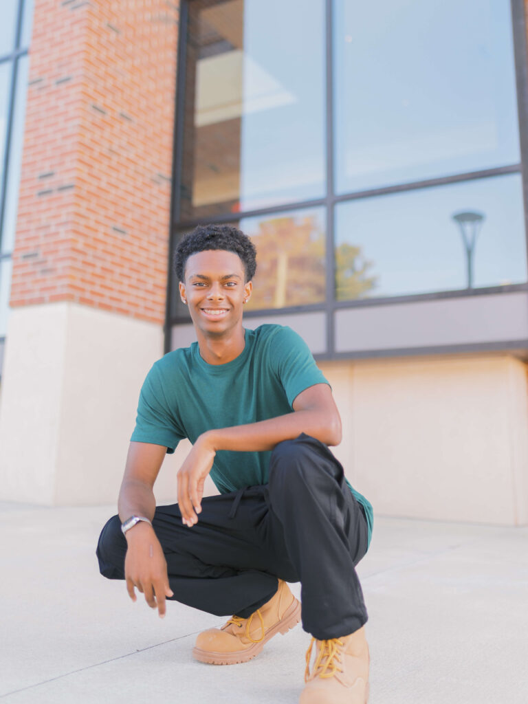 Class of 2025 male senior crouching in front of a modern glass building in Downtown Lawrenceville, Georgia, during a morning senior photo session.