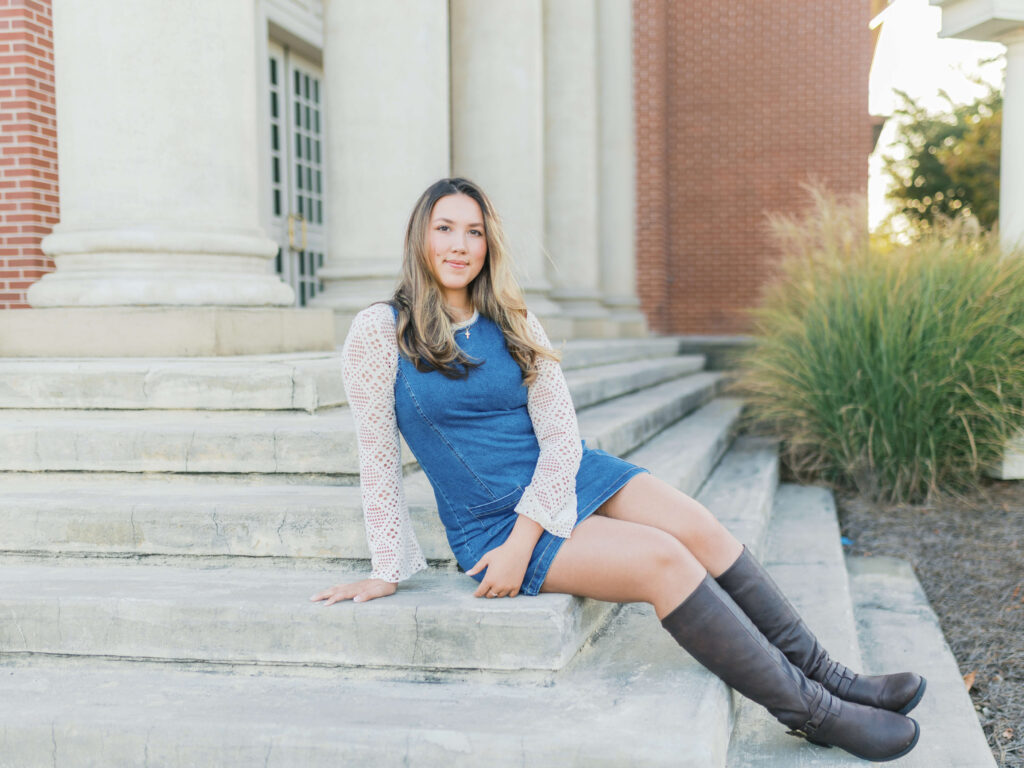 Senior girl from the Class of 2025 sitting on marble steps in Downtown Lawrenceville, Georgia, wearing a denim dress and knee-high boots during her morning portrait session.