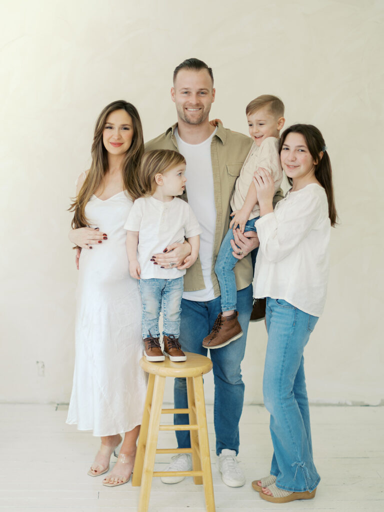 A smiling family of five poses for a portrait against a cream wall. The pregnant mother, father, and three children are dressed casually in light tops and denim.