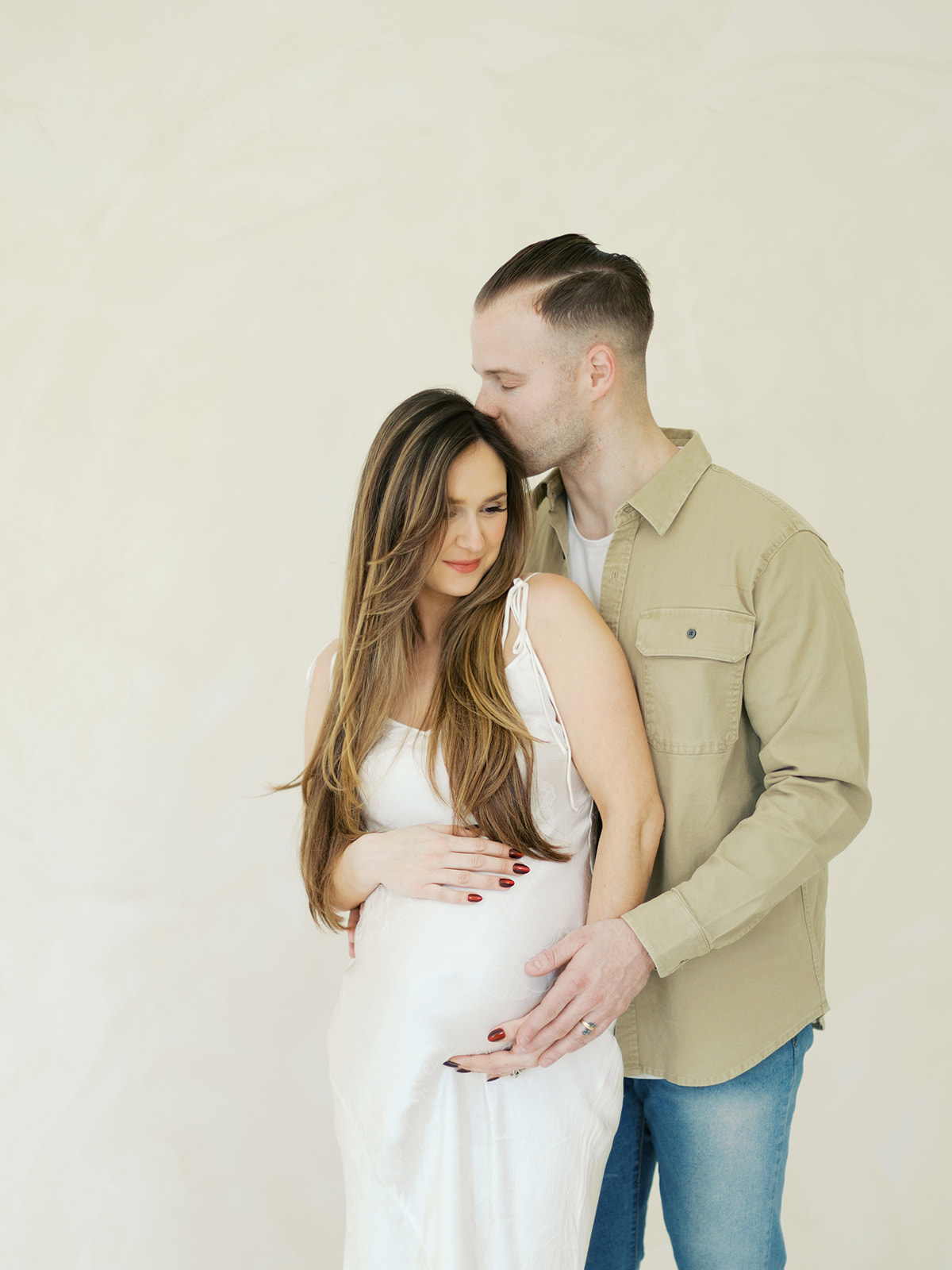 A man standing behind a pregnant woman, kissing the top of her head while they both rest their hands on her baby bump.